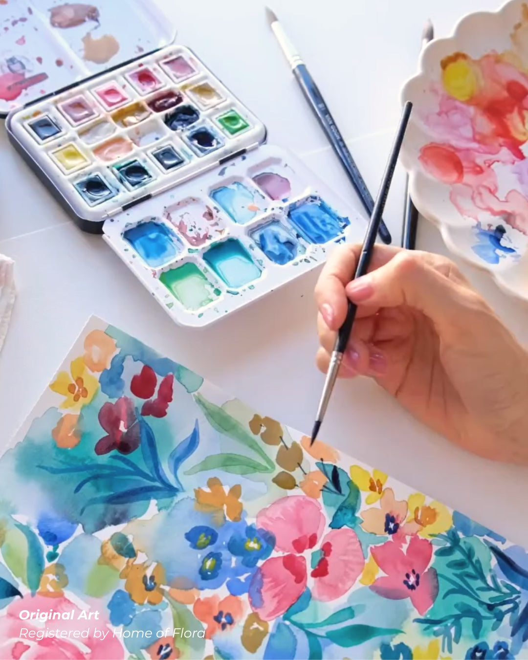 Hand holding a paintbrush over watercolor paintings with a watercolor set in the background.