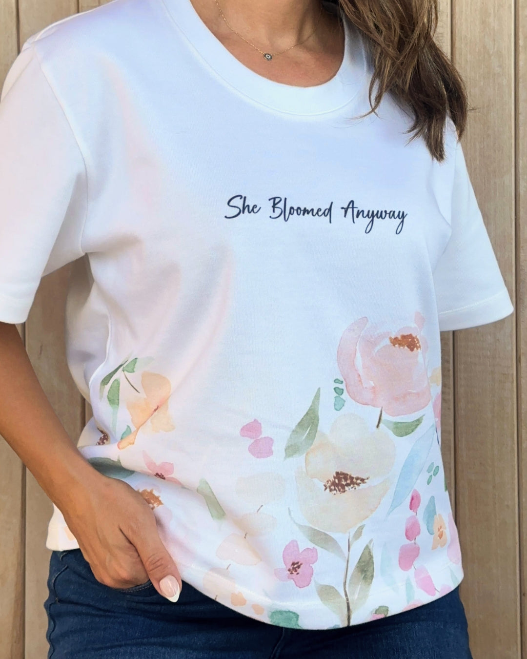 White t-shirt with floral design and 'She Bloomed Anyway' text worn by a person.