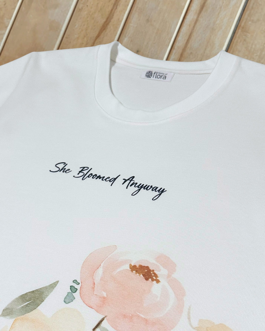 White t-shirt with floral design and text 'She Bloomed Anyway' on a wooden surface