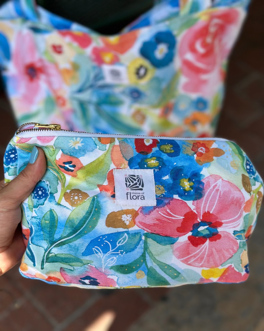 Floral-patterned pouch held by a hand with 'flora' brand label.