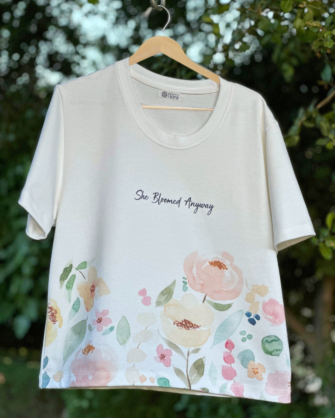 White t-shirt with floral design and text 'She Bloomed Anyway' on a hanger against a green leafy background.