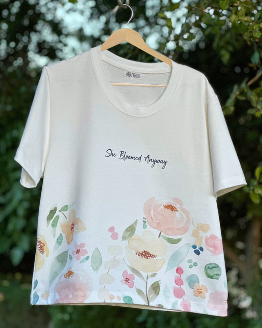 White t-shirt with floral design and text 'She Bloomed Anyway' on a hanger against a green leafy background.