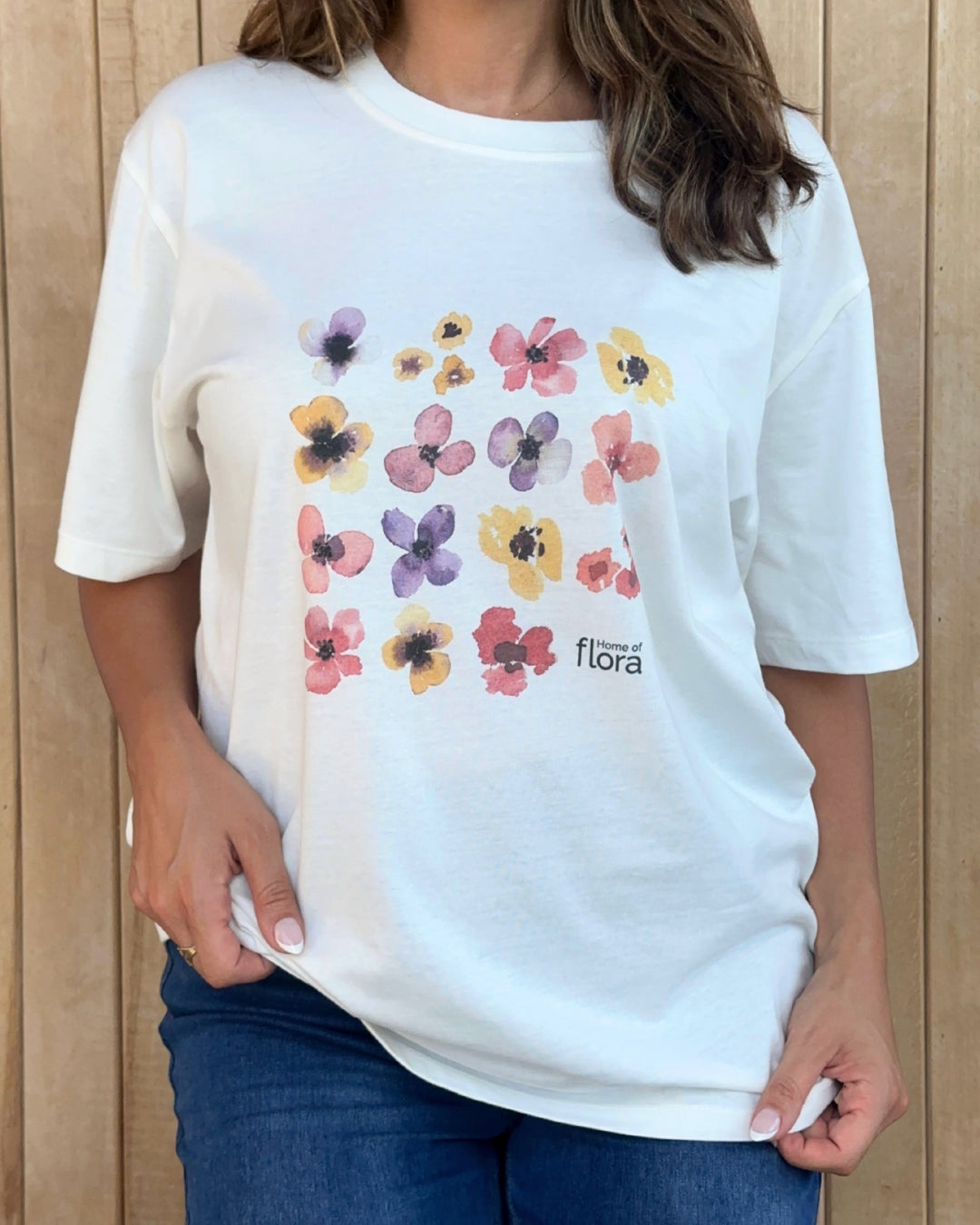 Person wearing a white t-shirt with colorful floral designs and 'Home of Flora' text against a wooden background.
