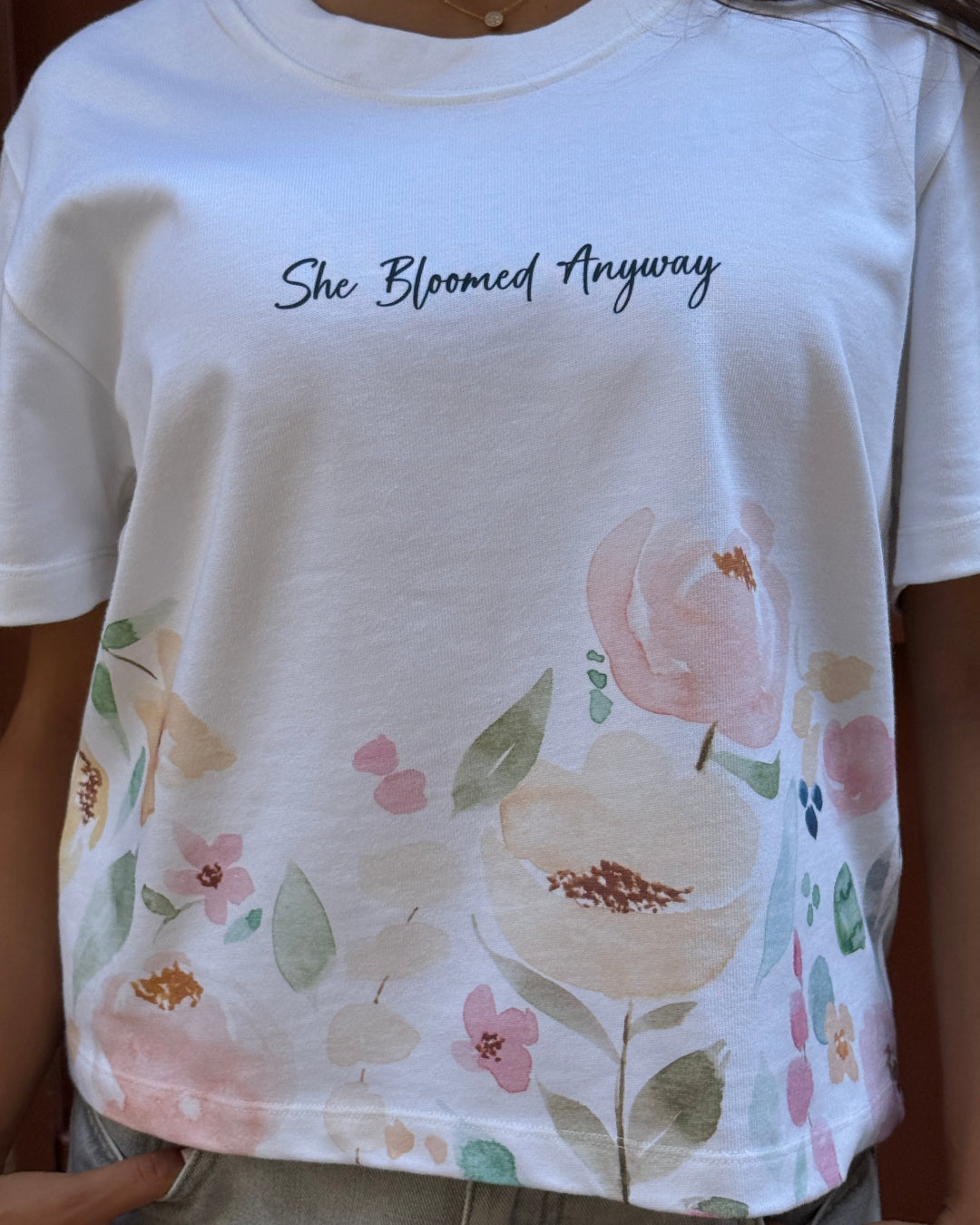 Person wearing a white t-shirt with floral design and 'She Bloomed Anyway' text.