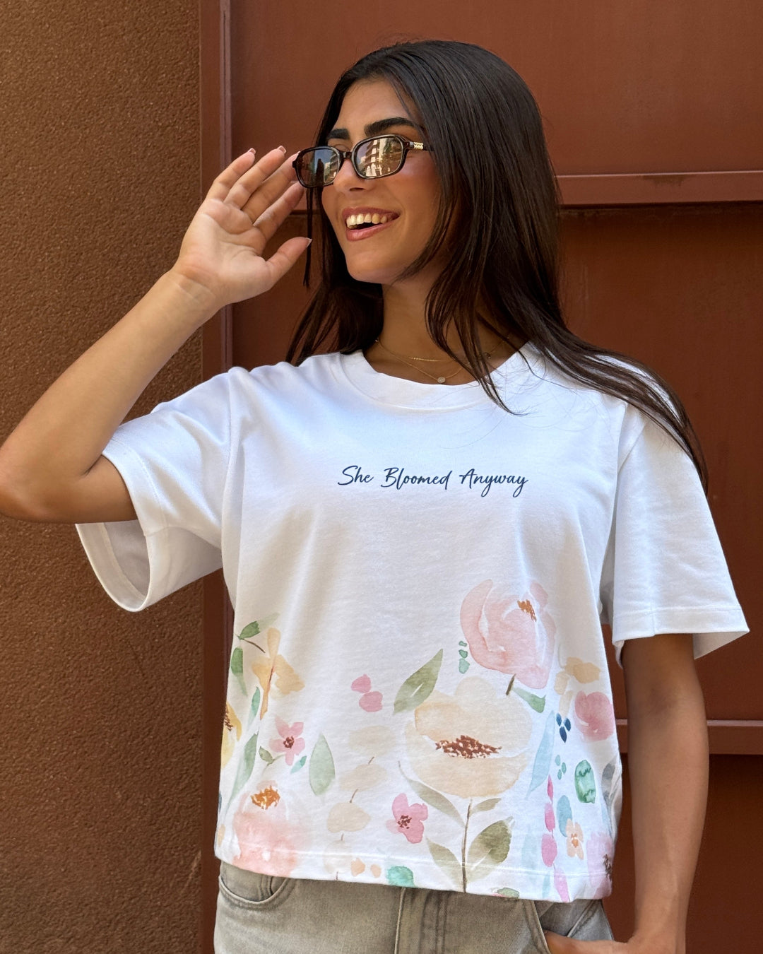 Woman wearing a white t-shirt with floral design and text, standing against a brown wall.