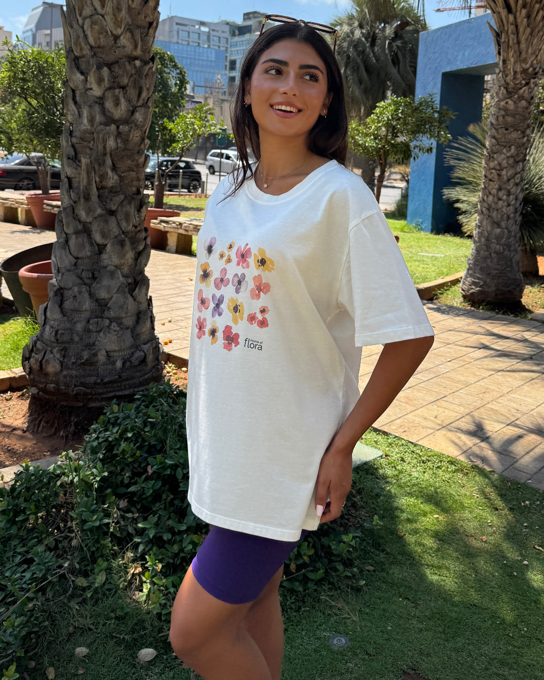 Woman wearing a white t-shirt with colorful graphics in an outdoor setting with palm trees and buildings.
