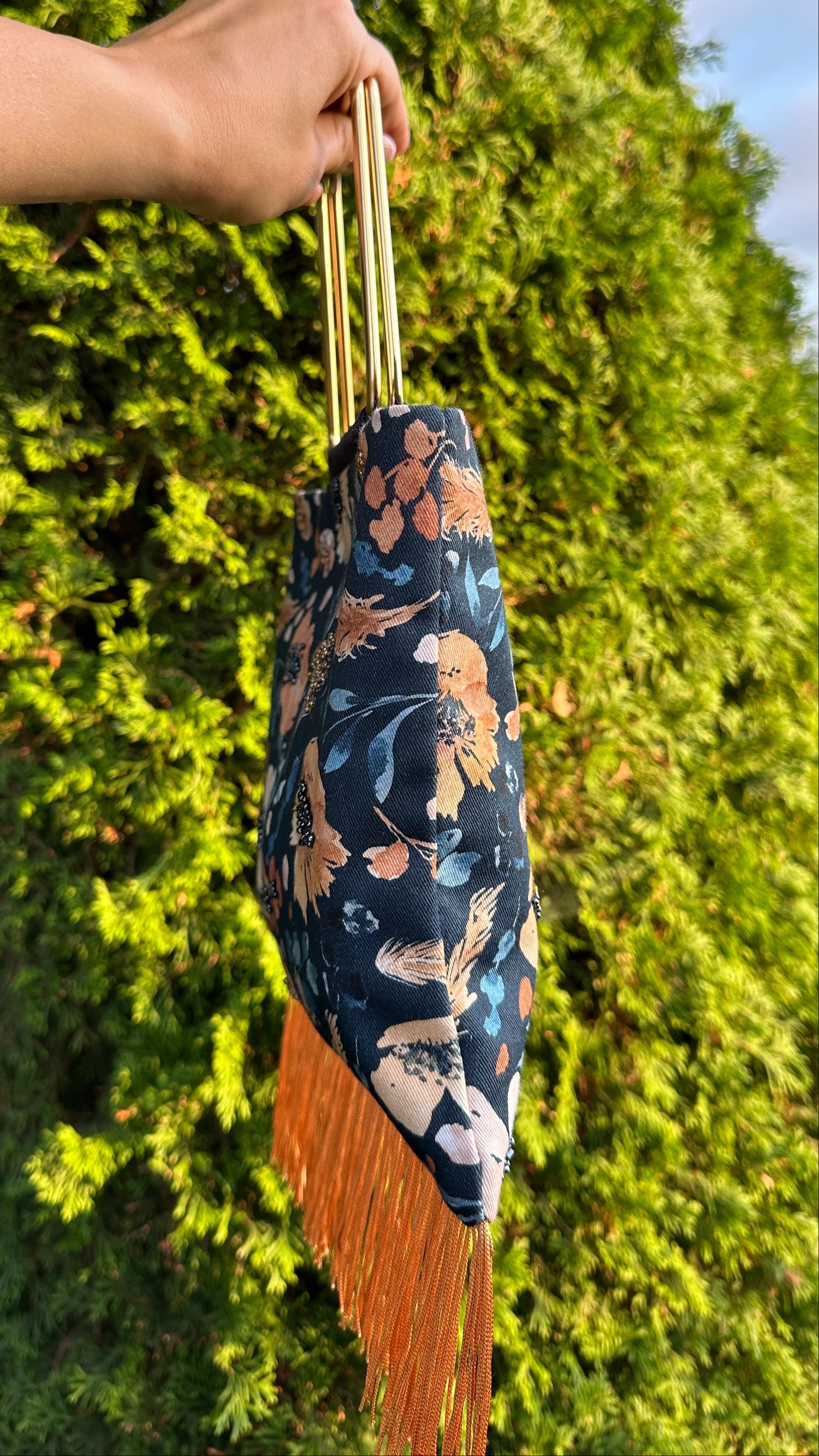 A hand holds a small fabric handbag with a dark blue floral print and long orange-brown fringe, set against a background of green foliage.