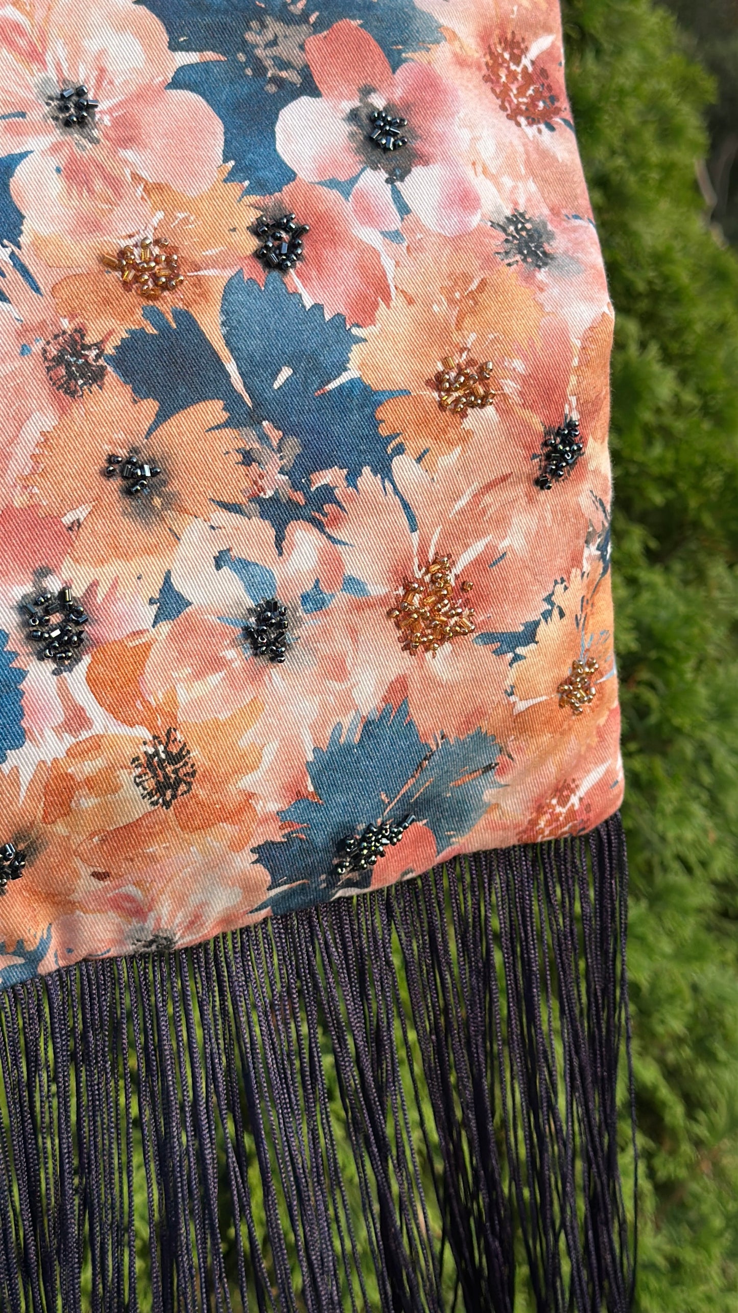 A close-up of a floral fabric with peach, pink, and blue watercolor-style flowers, black beaded centers, and long black fringe hanging from the bottom, set against a blurred green outdoor background.