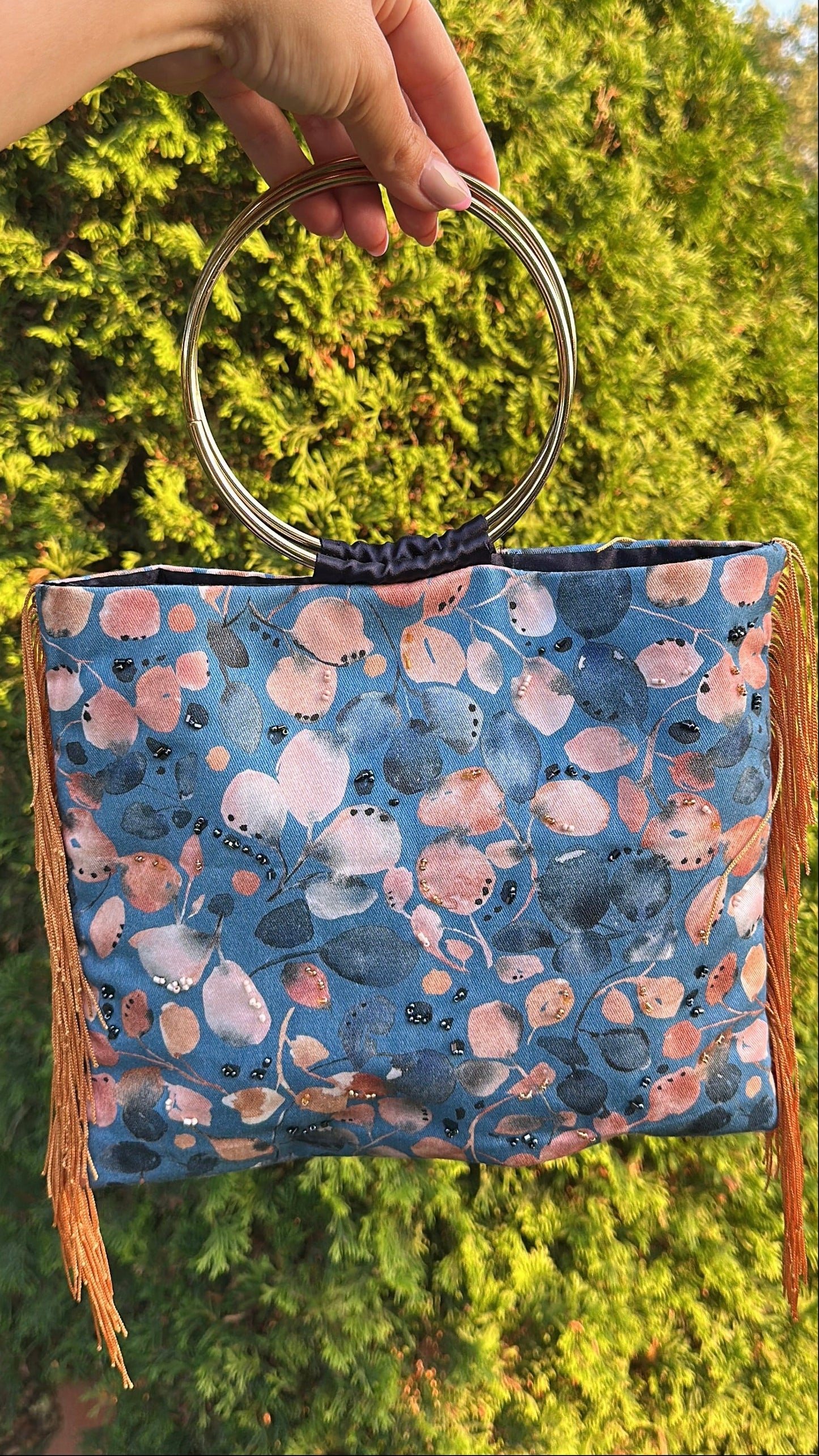 A hand holds a blue patterned handbag with a circular metal handle and brown fringe, set against a background of green foliage.