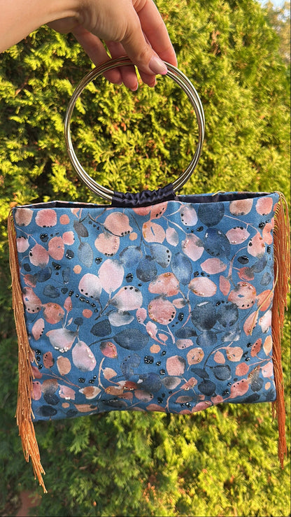 A hand holds a blue patterned handbag with a circular metal handle and brown fringe, set against a background of green foliage.