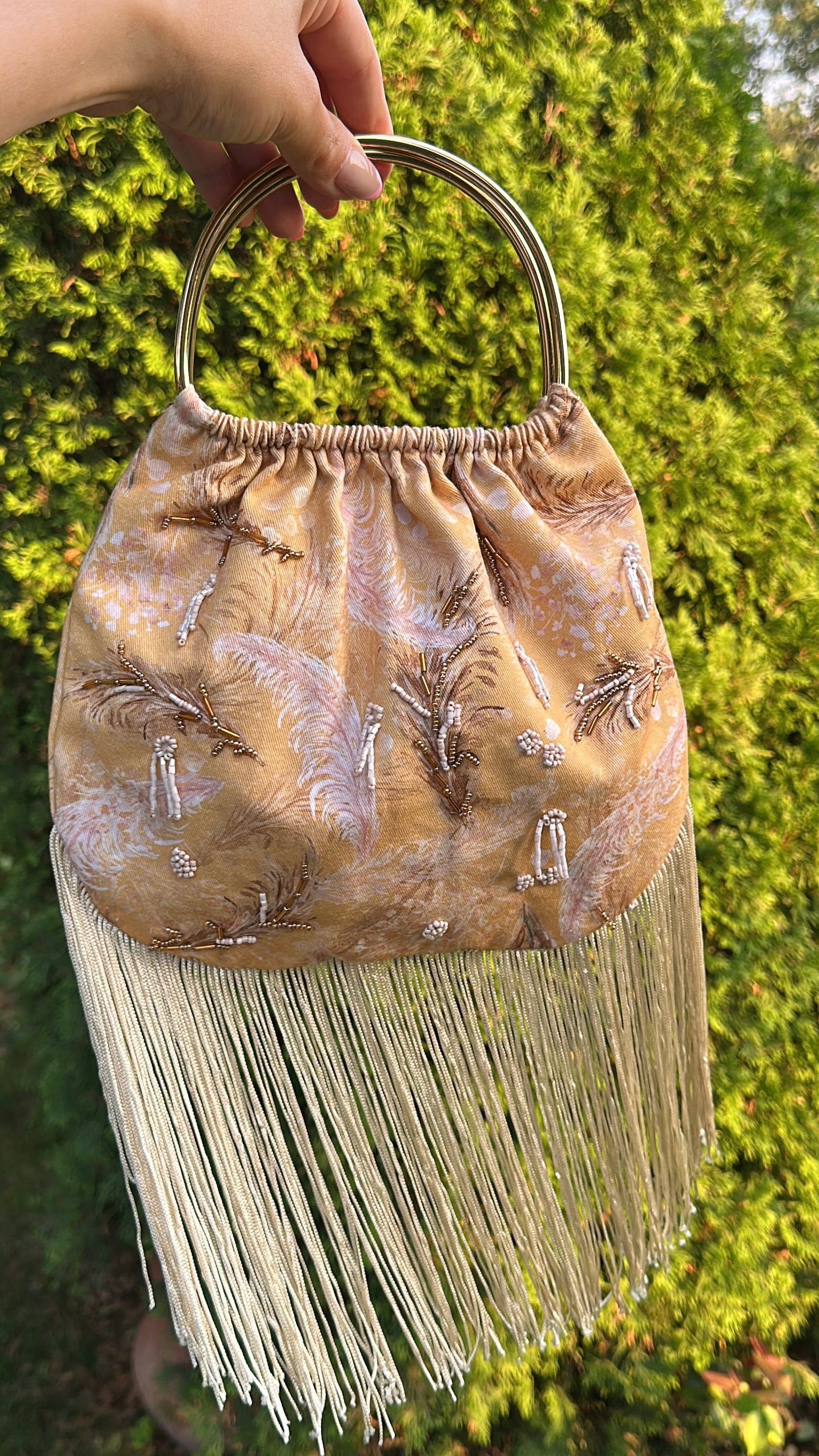 A hand holds a small beige fringed handbag with a circular metal top handle, featuring embroidered details, against a background of green foliage.