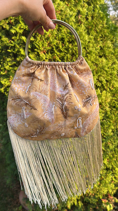 A hand holds a small beige fringed handbag with a circular metal top handle, featuring embroidered details, against a background of green foliage.