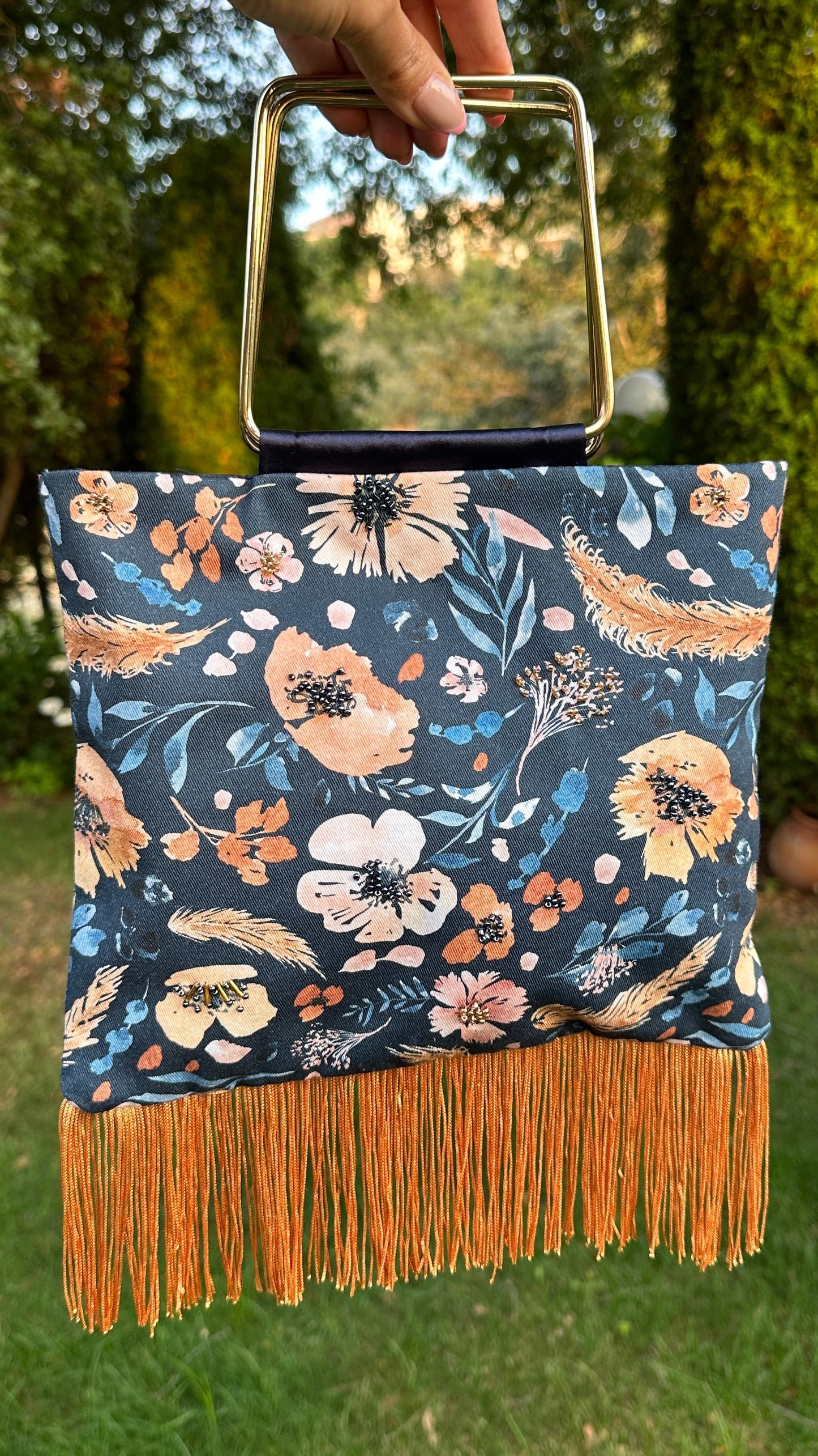 A hand holds a square floral clutch bag with a gold metal handle and orange fringe, set against a blurred green garden background.