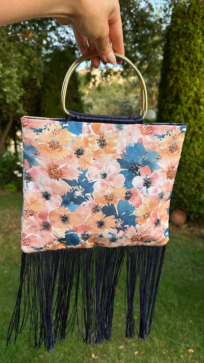 A hand holds a square floral handbag with a gold-tone circular handle and long dark fringe, photographed outdoors against green trees and grass.