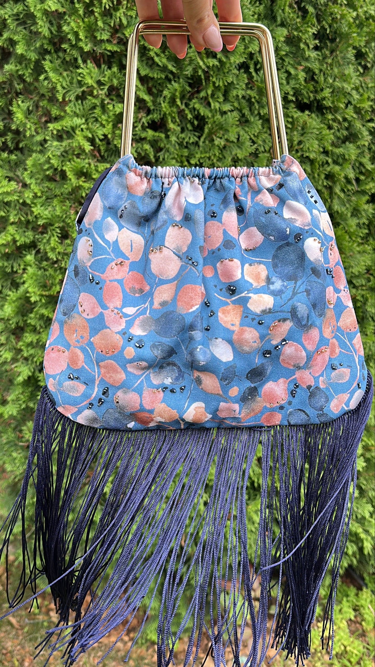 A hand holds a blue patterned fringe handbag with a gold-tone metal top handle against a green hedge background.