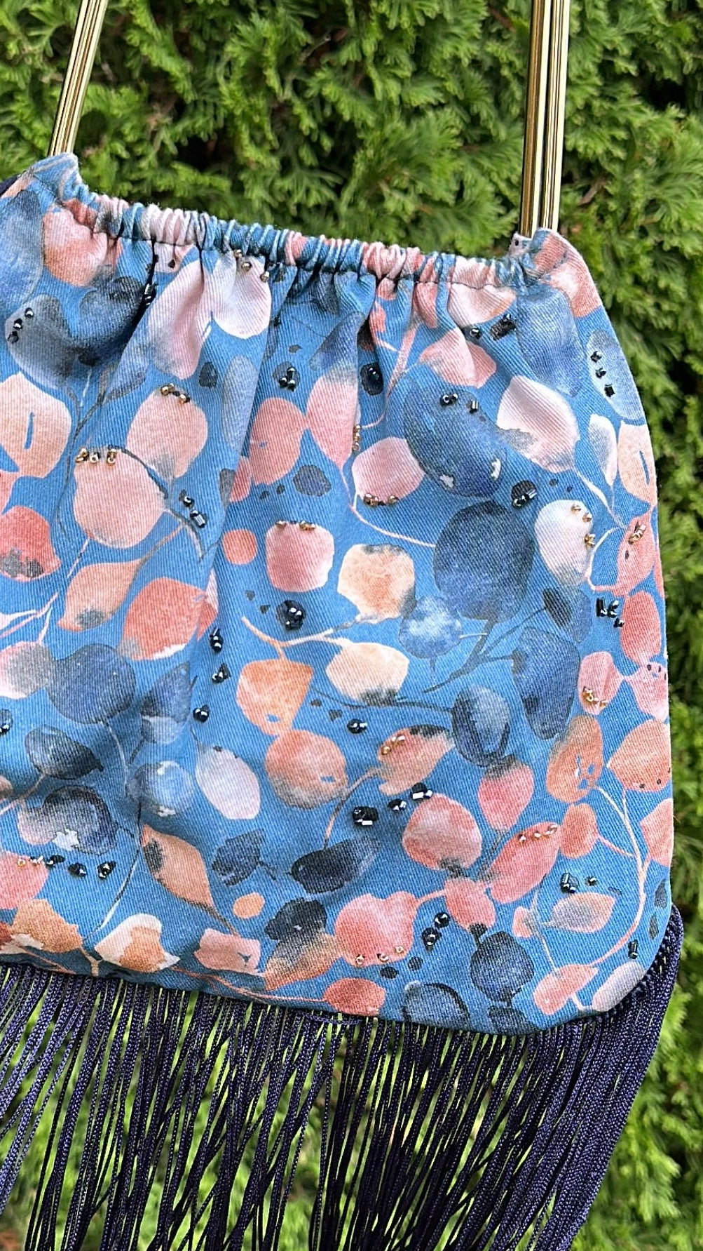 A hand holds a blue patterned fringe handbag with a gold metal top handle against a background of green hedges.