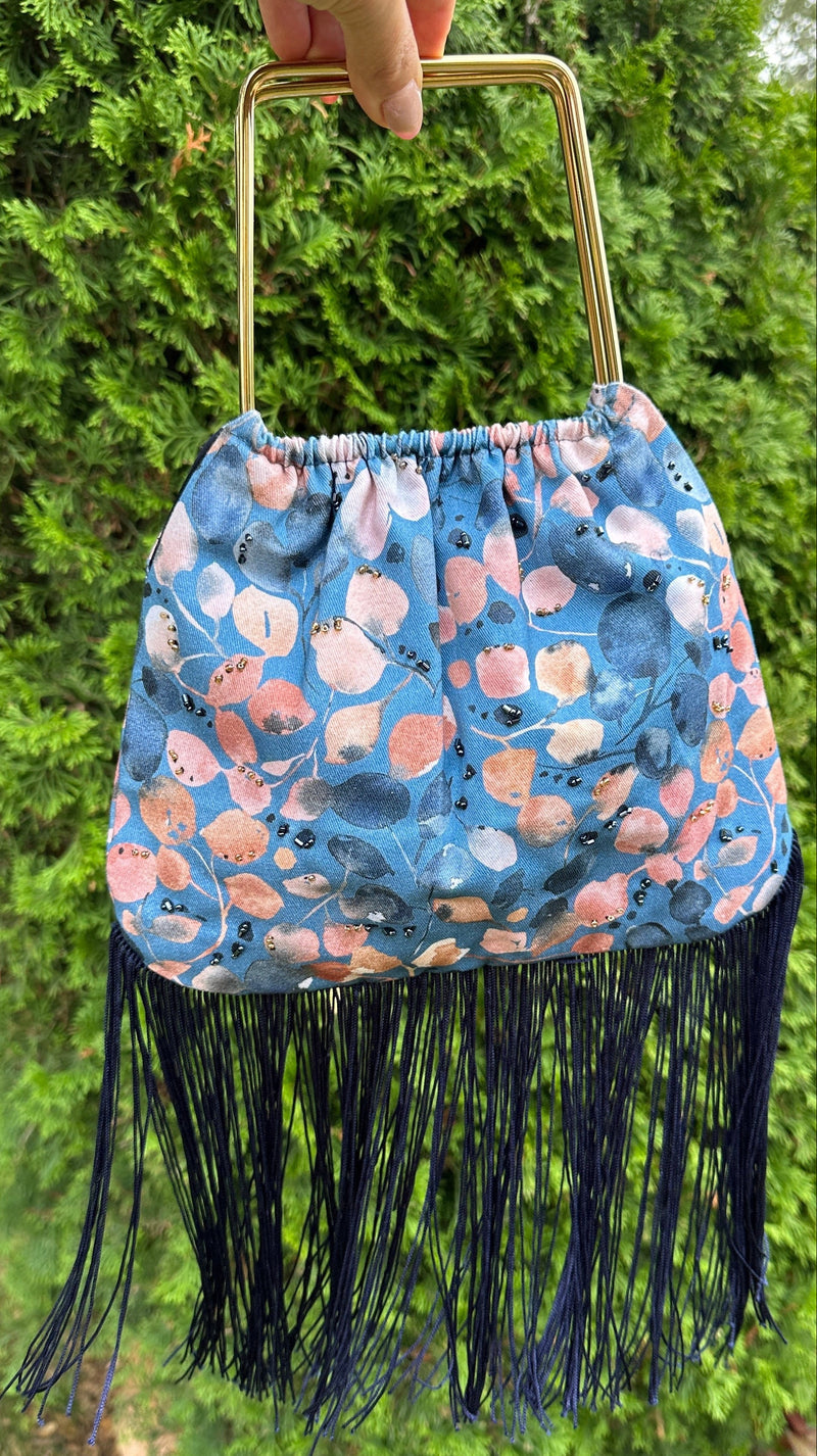 A blue patterned fringe handbag with a gold metal top handle, held by a hand against a background of green hedges.