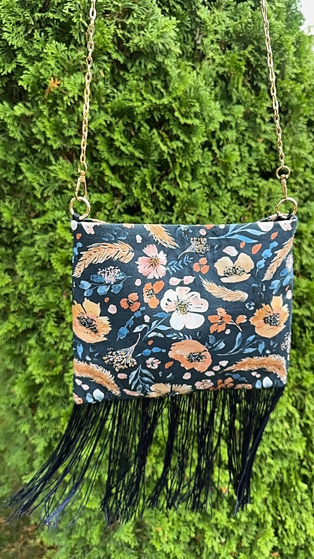 A small rectangular handbag with a dark navy floral print, black fringe trim, and a gold-tone chain strap, photographed outdoors against a green hedge background.
