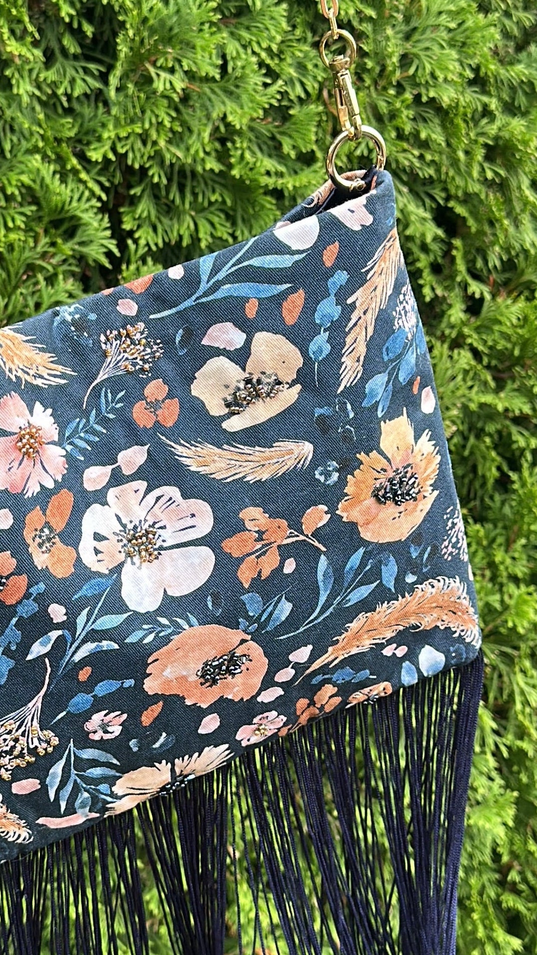 A navy blue floral fringe bag with a gold chain strap, hanging against a green hedge background.