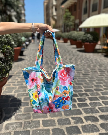 Colorful floral tote bag held by a person on a street with buildings and plants in the background