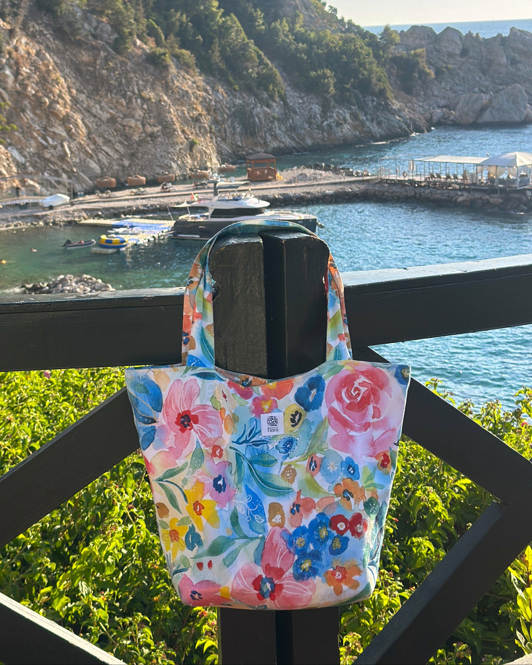 Floral tote bag hanging on a railing with a scenic background of water and mountains.